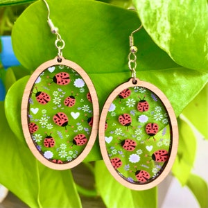 May include: A pair of oval-shaped wooden earrings with a clear resin insert featuring a repeating pattern of red ladybugs and white flowers on a green background.