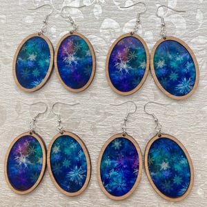 Pode incluir: Seis pares de brincos ovais de madeira com um design azul e roxo semelhante a uma gal&aacute;xia com flocos de neve brancos. Os brincos t&ecirc;m ganchos prateados.