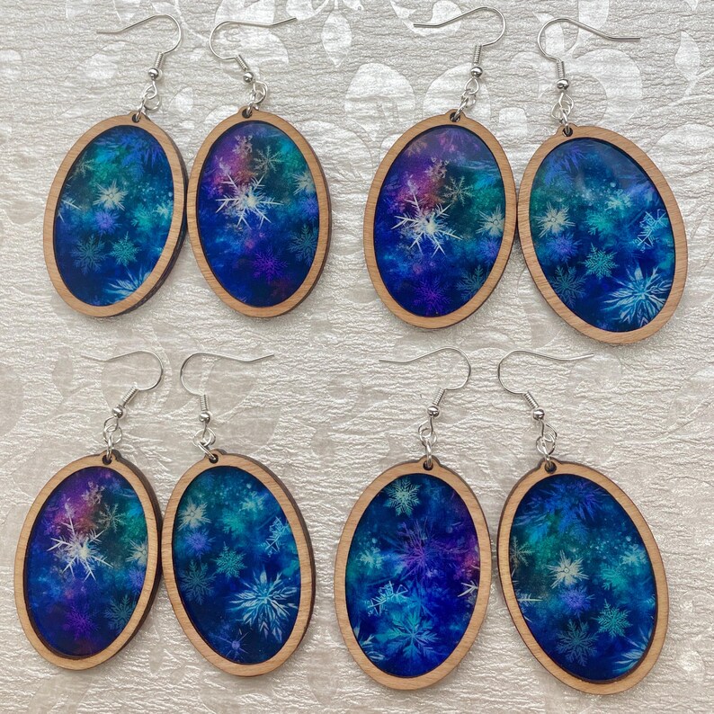Pode incluir: Seis pares de brincos ovais de madeira com um design azul e roxo semelhante a uma gal&aacute;xia com flocos de neve brancos. Os brincos t&ecirc;m ganchos prateados.