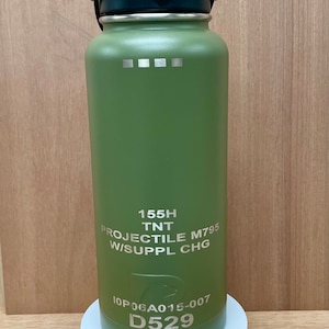 May include: A green metal water bottle with a black lid and a silver label that reads "155H TNT PROJECTILE M795 W/SUPPL CHG 10P06A015-007 D529".