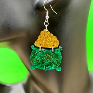 May include: Green and gold glitter pot of gold earrings. The earrings are shaped like a pot with a lid full of gold coins.