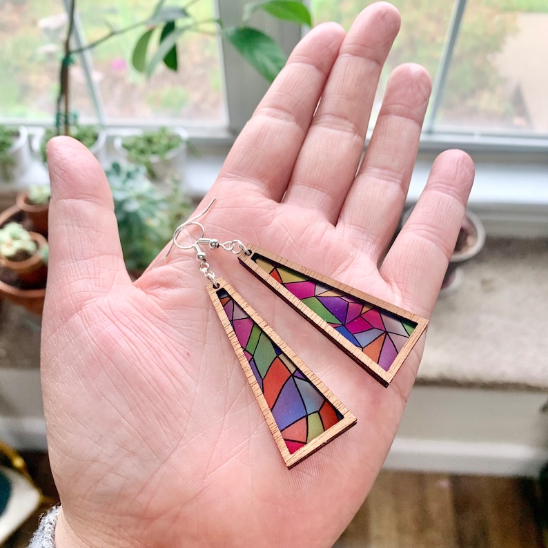 May include: A pair of wooden triangle earrings with a stained glass design featuring colorful geometric shapes. The earrings are made of natural wood and have silver hooks.
