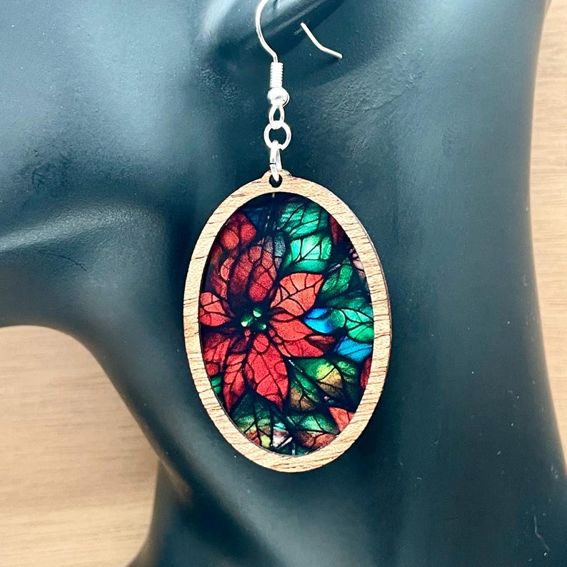 May include: Oval wooden earrings featuring a stained glass style poinsettia design. The earrings are red, green and blue.