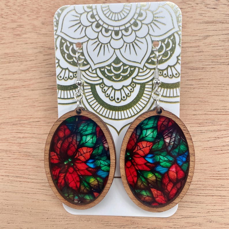 May include: A pair of oval-shaped wooden earrings with a stained glass design of red poinsettia flowers and green leaves. The earrings have silver hooks.