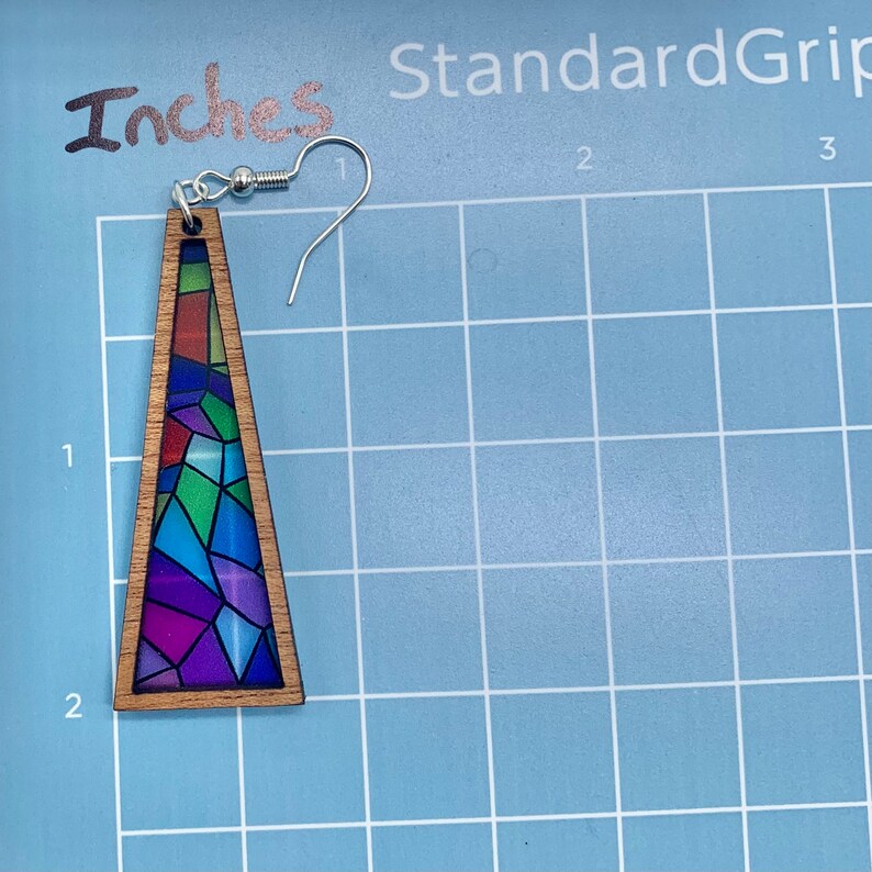 May include: A pair of wooden triangle earrings with a stained glass design featuring a variety of colors, including blue, green, purple, and red. The earrings are approximately 2 inches long.