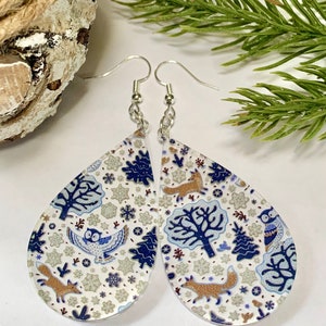 May include: Teardrop shaped earrings with a white background and a blue and gold pattern of owls, foxes, trees, and snowflakes. The earrings are made of clear acrylic and have silver hooks.