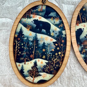 Pu&ograve; includere: Orecchini ovali in legno con una scena di foresta invernale. La scena rappresenta due orsi neri che camminano attraverso una foresta innevata con pini e una capanna sullo sfondo. La scena &egrave; circondata da una cornice in legno.