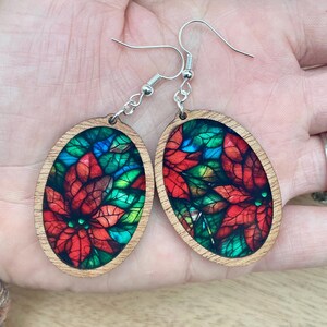 May include: Oval-shaped wooden earrings with a stained glass design featuring red poinsettia flowers and green leaves. The earrings have silver hooks.