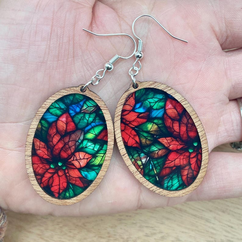 May include: Oval-shaped wooden earrings with a stained glass design featuring red poinsettia flowers and green leaves. The earrings have silver hooks.
