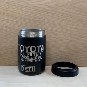 May include: A black YETI tumbler with a silver rim and a black rubber base. The tumbler features the Toyota logo and "OIL FILTER 15600-41010" printed in white. The text "MADE IN JAPAN" and Japanese characters are also visible.