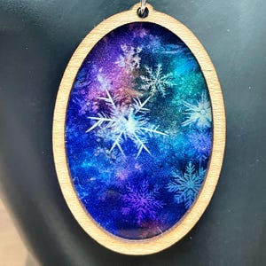 Pode incluir: Pingente oval de madeira com um inserto de resina transparente que apresenta um fundo de gal&aacute;xia azul, roxo e verde com desenhos de flocos de neve brancos.
