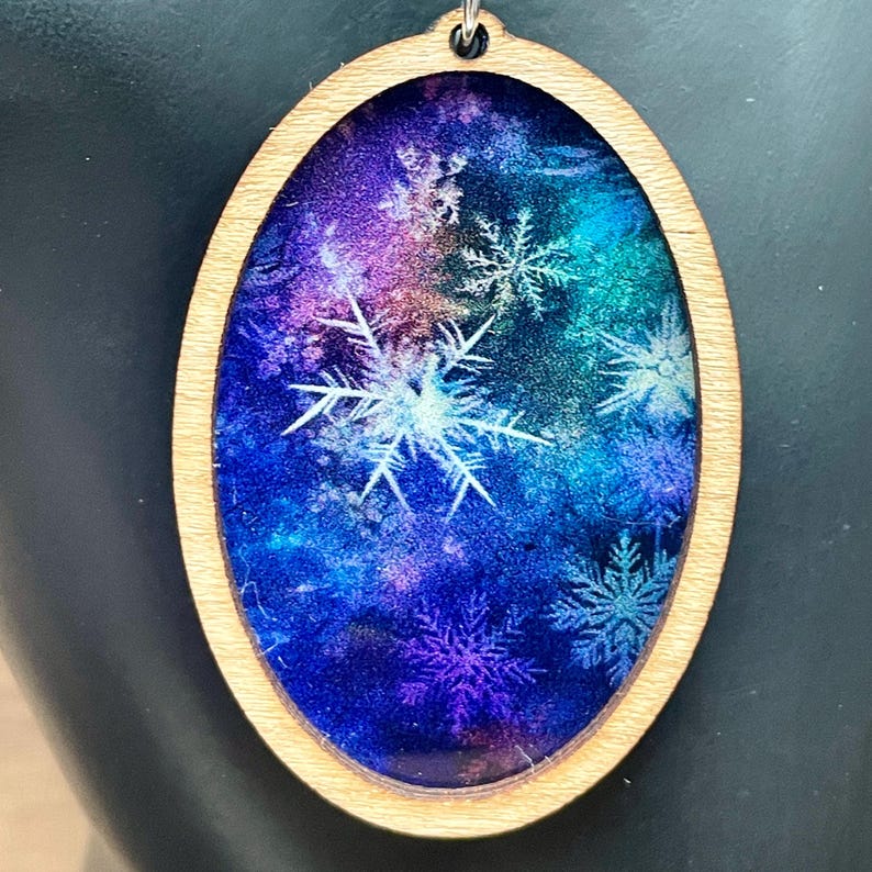 Pode incluir: Pingente oval de madeira com um inserto de resina transparente que apresenta um fundo de gal&aacute;xia azul, roxo e verde com desenhos de flocos de neve brancos.