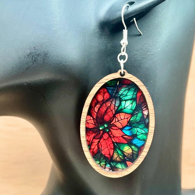May include: Oval wooden earrings featuring a stained glass style poinsettia design. The earrings are red, green and blue.