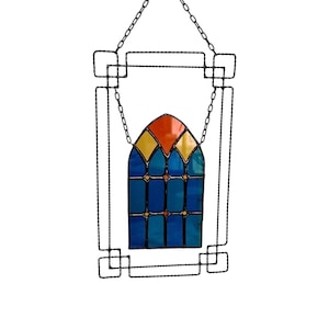 Könnte beinhalten: Dekoration für ein Fenster aus Buntglas. Das Fenster hat ein farbenfrohes Design mit blauen, gelben und orangen Glasscheiben. Es wird von einer schwarzen Metallstruktur umrahmt und an einer Kette aufgehängt.