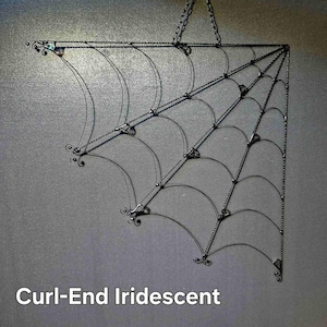 Puede incluir: Una telaraña decorativa de metal en forma de esquina, con una cadena para colgar. La telaraña presenta líneas curvas y pequeños remolinos decorativos en los extremos. El texto "Curl-End Iridescent" se muestra en la parte inferior.
