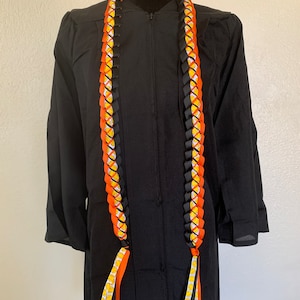 May include: A black graduation gown with a yellow, orange, and black lei. The lei has a pattern of small yellow circles on the orange ribbon.