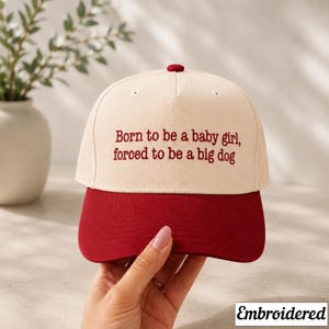 May include: A baseball cap with a cream-colored crown and a red brim. The cap features the embroidered text: "Born to be a baby girl, forced to be a big dog." A small red button is on top. The word "Embroidered" is in the bottom right corner.