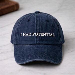 Pode incluir: Boné de beisebol azul marinho com o texto branco "I HAD POTENTIAL" na frente. O boné tem uma aba curva e um pequeno botão na parte superior. O chapéu é feito de um tecido macio e texturizado.