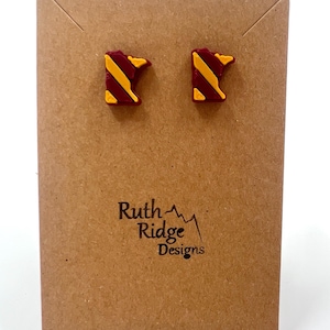 May include: A pair of stud earrings shaped like the state of Minnesota, with a maroon and gold design. The earrings are displayed on a brown card with the text "Ruth Ridge Designs".