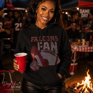 May include: Black hooded sweatshirt with the text "FALCONS FAN" in red and silver rhinestones. The design includes a high heel shoe and a football, also in rhinestones. The person is holding a red cup.
