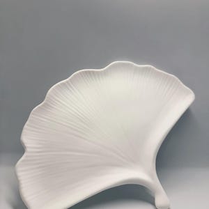 Puede incluir: Un plato decorativo blanco con forma de hoja. El plato tiene una superficie detallada y texturizada con un patrón en forma de abanico. El diseño es una representación estilizada de una hoja de ginkgo. El plato es de cerámica o porcelana.