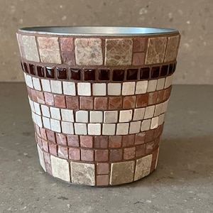 Può includere: Un vaso rotondo in terracotta con un design a mosaico. Il mosaico è fatto di piccole piastrelle quadrate in tonalità di marrone, rosa e bianco. Il vaso ha un bordo argentato.