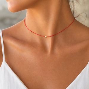 Puede incluir: Un collar tipo gargantilla minimalista que presenta un cordón rojo fino y vibrante con una única cuenta esférica pequeña de tono dorado pulido centrada en la parte delantera. El cordón se ajusta al cuello, ofreciendo una estética limpia y sencilla.