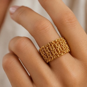 Puede incluir: Un anillo ancho de color dorado que presenta un patrón tejido detallado al estilo de una cota de malla. La banda está construida con numerosos eslabones metálicos pequeños entrelazados que crean un diseño texturizado y calado.