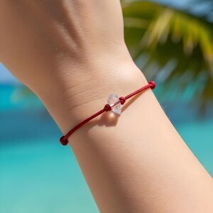 May include: A red string bracelet with a clear, faceted crystal bead. The bracelet is tied with knots and is worn on a wrist. The background shows a blurred view of a tropical beach with palm trees and blue water.