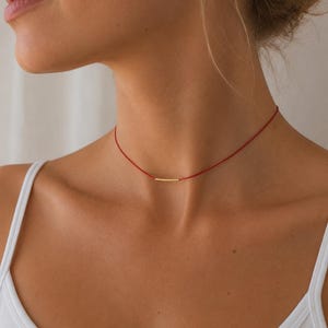 Puede incluir: Un collar tipo gargantilla minimalista que presenta un cordón fino de color rojo vibrante y una pequeña barra horizontal de metal dorado con una superficie texturizada. El delicado cordón rodea el cuello, resaltando la pieza metálica central.