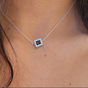 Puede incluir: Un collar de cadena de plata con un colgante cuadrado que presenta una pequeña piedra negra en el centro.