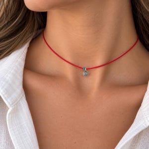 Puede incluir: Un collar de cordón rojo fino con un pequeño colgante circular de tono plateado con un diseño de espiral grabado. El colgante está decorado con una diminuta cuenta de color turquesa situada justo al lado del dije plateado en el cordón.