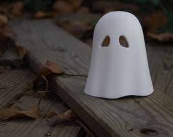 May include: A small, white ceramic ghost figurine with two eye holes. The ghost is sitting on a weathered wooden surface, with dried leaves scattered around. The figurine has a simple, rounded design, perfect for Halloween decor.