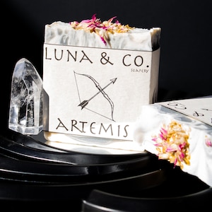 May include: A white soap bar with a grey marbled design and pink flower petals on top. The soap bar is labelled "LUNA & CO. SOAPERY" and "ARTEMIS" with a black and white illustration of a bow and arrow.