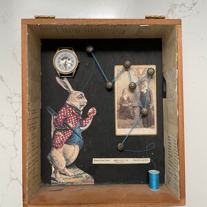 Könnte beinhalten: Eine Holzkiste mit schwarzem Hintergrund enthält eine Vintage-Illustration eines Kaninchens, das eine Weste trägt und eine Spielkarte hält. Eine kleine Uhr und ein Foto von zwei Frauen sind ebenfalls in der Kiste enthalten. Die Kiste ist mit blauem Faden und Messingnägeln verziert.