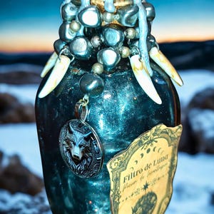 May include: A teal-colored, decorative glass bottle with a silver wolf pendant and a label that reads "Filtro de Luna". The bottle is adorned with beads, decorative accents, and what appear to be animal teeth. The background shows a night sky with a full moon.