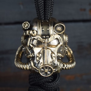 May include: A gold-colored metal charm in the shape of a Fallout Power Armor helmet. The charm is attached to a black paracord lanyard.