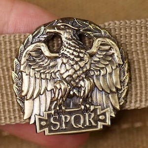 May include: A bronze-coloured metal emblem featuring a detailed eagle design with outstretched wings, surrounded by laurel leaves. The emblem is affixed to a tan fabric strap. The letters "SPQR" are inscribed on a rectangular banner below the eagle.