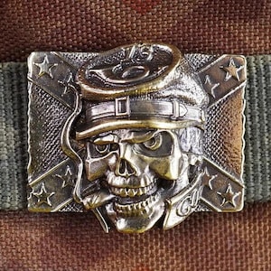 以下が含まれることがあります： 迷彩柄の背景に、帽子をかぶった骸骨が特徴的な、アンティークスタイルのベルトバックル。 バックルは金属製で、星や旗のようなデザインなど、複雑なディテールが施されています。 ベルトはオリーブグリーンです。