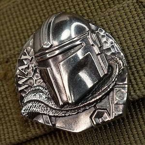 Op de afbeelding: Een zilverkleurige Mandalorian helmspeld met ingewikkelde details. De speld heeft de tekst "WAY" en is geplaatst tegen een getextureerde olijfgroene achtergrond. Het woord "Melchior" staat rechtsboven.