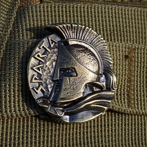 May include: A bronze-colored metal pin with a Spartan helmet design and the word "SPARTA" inscribed around the edge. The pin is attached to a green fabric background.