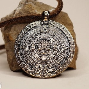May include: A gold-toned pendant necklace featuring a detailed replica of the Aztec calendar stone. The pendant is round and has a brown leather cord.