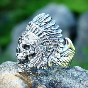 Op de afbeelding: Een zilveren ring met een gedetailleerd schedelontwerp dat een hoofdtooi met veren draagt. De schedel heeft een wijd open mond en de hoofdtooi heeft vleugels aan de zijkant.