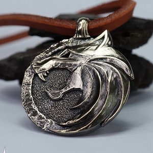May include: A brass pendant featuring a detailed wolf's head design within a circular frame, attached to a brown leather cord. The pendant has a textured surface and is set against a dark background. The word "Brass" is visible in the upper right corner.