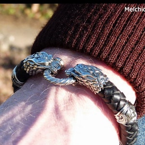 Könnte beinhalten: Ein schwarzes Lederarmband mit silbernen Wolfs-Kopf-Verschlüssen. Das Armband ist geflochten und hat ein detailliertes Design.