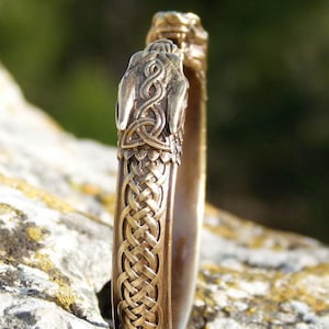 Può includere: Un bracciale in ottone con nodi celtici e dettagli di testa di drago. Il bracciale presenta un design intricato con motivi intrecciati e una finitura strutturata. La parola "Brass" è visibile in alto a destra.