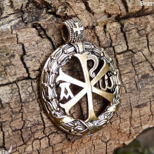 May include: A brass pendant with a wreath design and a cross with the letters 'A', 'W', and 'Ω' inside. The pendant is on a wooden surface.