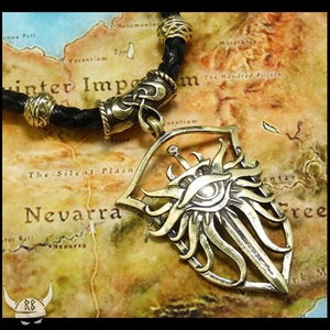 May include: A gold-toned necklace with a leather cord and a pendant featuring a stylized eye with a sword and sunburst design.