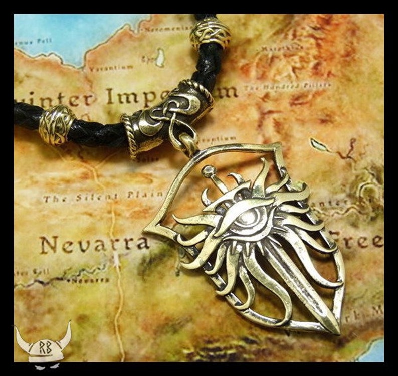 May include: A gold-toned necklace with a leather cord and a pendant featuring a stylized eye with a sword and sunburst design.