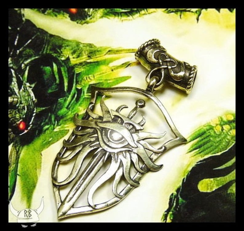 May include: A silver pendant in the shape of a shield with a sword and an eye inside. The pendant is on a green and brown background.